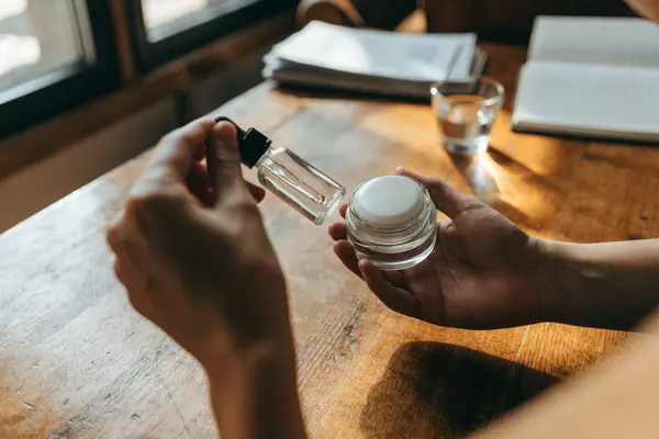 Primer plano con ángulo alto de manos sosteniendo un frasco gotero de vidrio abierto sin marca y un pequeño tarro de cosméticos en blanco sin marca sobre una mesa rústica de madera. Luz matutina suave. Primer plano con ángulo alto de manos sosteniendo un frasco gotero de vidrio abierto sin marca y un pequeño tarro de cosméticos en blanco sin marca sobre una mesa rústica de madera. Luz matutina suave.