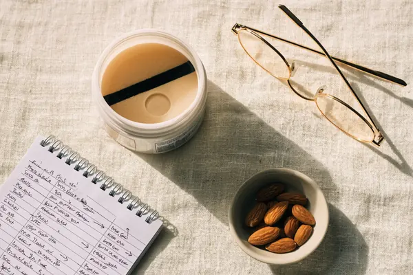 Vista cenital de un recipiente de cuidado de la piel abierto, gafas de lectura, un cuaderno con notas analíticas manuscritas y un cuenco de almendras sobre un mantel de lino texturizado.