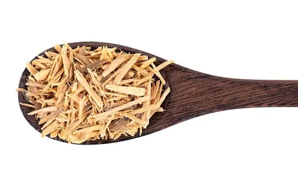 Naturaleza muerta macro con ingredientes botánicos secos y una cápsula blanca sobre una superficie de madera texturizada.
