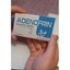 Un hombre argentino en un mostrador de farmacia comparando una caja pequeña de suplementos con su teléfono, sugiriendo que está comprobando si Adenofrin está disponible en las farmacias.