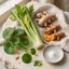 Bodegón editorial con hojas frescas de centella asiática, apio, raíz de jengibre, cúrcuma y una referencia a la vitamina C.