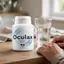 Paquete de Oculax junto a un vaso de agua sobre una mesa, con manos de adulto presentes.